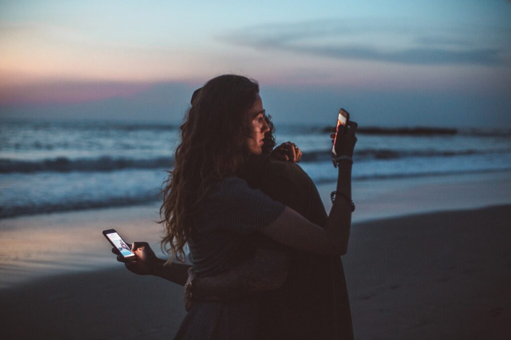 The impact of analog communication on relationships- Couple hugging on beach and using smartphone.jpg